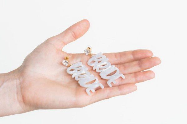 Bride earrings paired with a gold-capped champagne bottle, featuring elegant white script, ideal for a bridal shower and beautifully placed on a white fabric background.