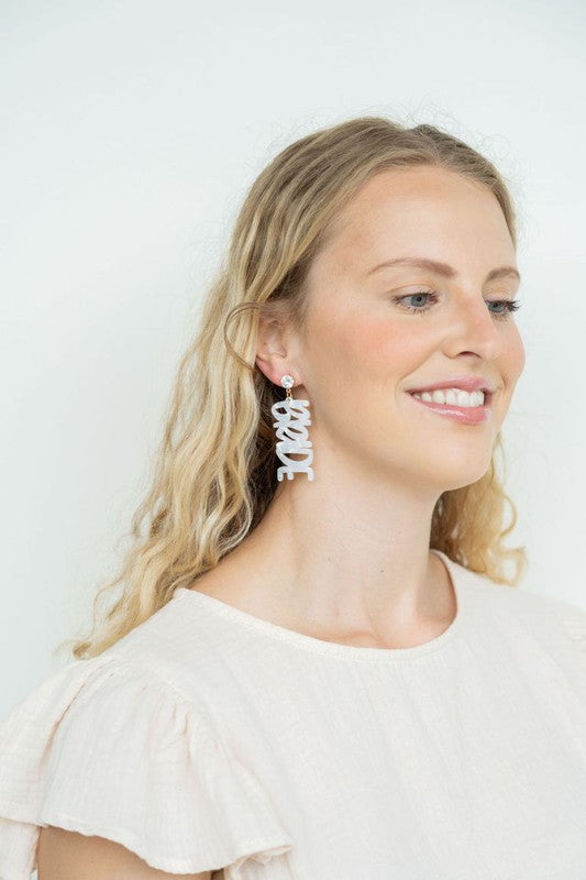 Bride earrings paired with a gold-capped champagne bottle, featuring elegant white script, ideal for a bridal shower and beautifully placed on a white fabric background.