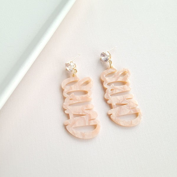 Two dangling earrings featuring "chaos" in peach-colored material, with exquisite hand-lettered calligraphy, are attached to gold posts adorned with clear stone accents. Perfectly named the "Bachelorette Squad," these earrings make a stunning bridal party gift and rest elegantly on a white surface.