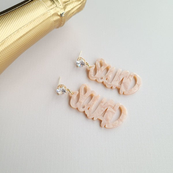 Two dangling earrings featuring "chaos" in peach-colored material, with exquisite hand-lettered calligraphy, are attached to gold posts adorned with clear stone accents. Perfectly named the "Bachelorette Squad," these earrings make a stunning bridal party gift and rest elegantly on a white surface.