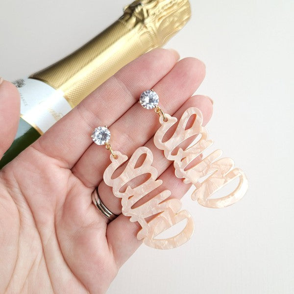 Two dangling earrings featuring "chaos" in peach-colored material, with exquisite hand-lettered calligraphy, are attached to gold posts adorned with clear stone accents. Perfectly named the "Bachelorette Squad," these earrings make a stunning bridal party gift and rest elegantly on a white surface.