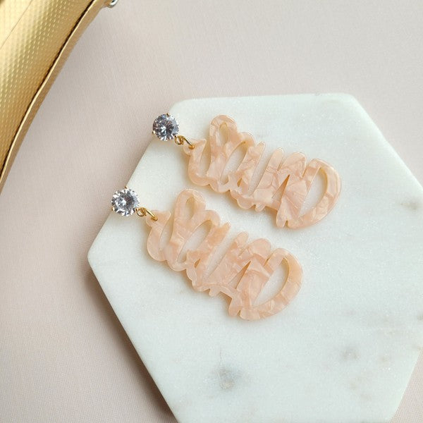 Two dangling earrings featuring "chaos" in peach-colored material, with exquisite hand-lettered calligraphy, are attached to gold posts adorned with clear stone accents. Perfectly named the "Bachelorette Squad," these earrings make a stunning bridal party gift and rest elegantly on a white surface.