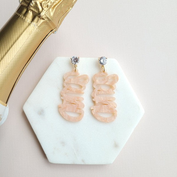 Two dangling earrings featuring "chaos" in peach-colored material, with exquisite hand-lettered calligraphy, are attached to gold posts adorned with clear stone accents. Perfectly named the "Bachelorette Squad," these earrings make a stunning bridal party gift and rest elegantly on a white surface.