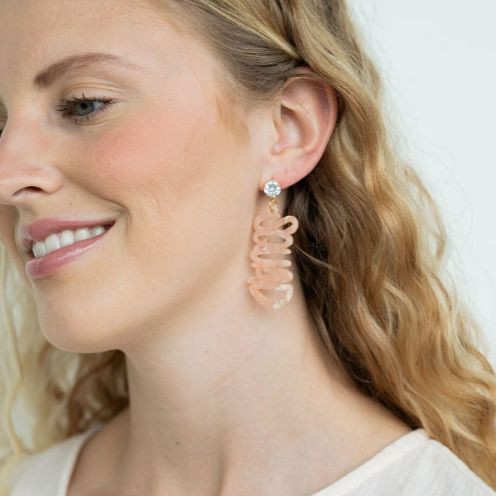 Two dangling earrings featuring "chaos" in peach-colored material, with exquisite hand-lettered calligraphy, are attached to gold posts adorned with clear stone accents. Perfectly named the "Bachelorette Squad," these earrings make a stunning bridal party gift and rest elegantly on a white surface.