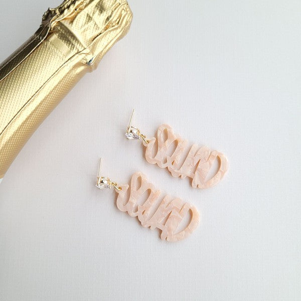 Two dangling earrings featuring "chaos" in peach-colored material, with exquisite hand-lettered calligraphy, are attached to gold posts adorned with clear stone accents. Perfectly named the "Bachelorette Squad," these earrings make a stunning bridal party gift and rest elegantly on a white surface.