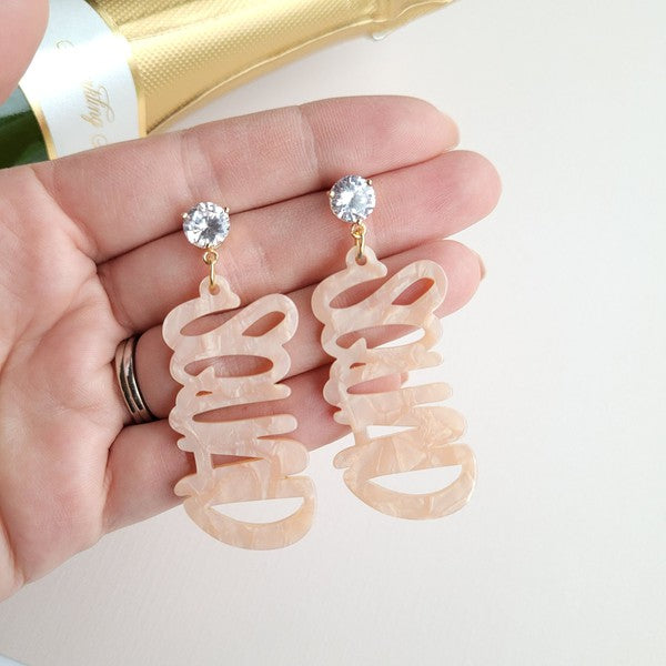 Two dangling earrings featuring "chaos" in peach-colored material, with exquisite hand-lettered calligraphy, are attached to gold posts adorned with clear stone accents. Perfectly named the "Bachelorette Squad," these earrings make a stunning bridal party gift and rest elegantly on a white surface.