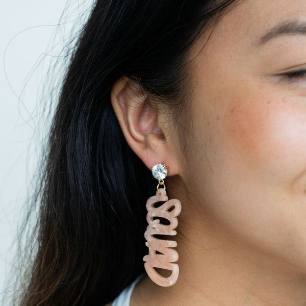 Two dangling earrings featuring "chaos" in peach-colored material, with exquisite hand-lettered calligraphy, are attached to gold posts adorned with clear stone accents. Perfectly named the "Bachelorette Squad," these earrings make a stunning bridal party gift and rest elegantly on a white surface.