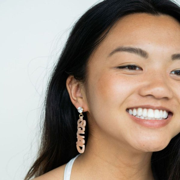 Two dangling earrings featuring "chaos" in peach-colored material, with exquisite hand-lettered calligraphy, are attached to gold posts adorned with clear stone accents. Perfectly named the "Bachelorette Squad," these earrings make a stunning bridal party gift and rest elegantly on a white surface.