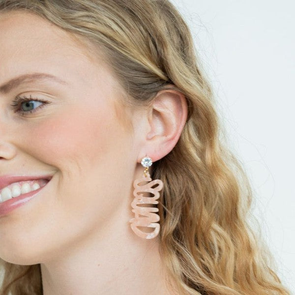 Two dangling earrings featuring "chaos" in peach-colored material, with exquisite hand-lettered calligraphy, are attached to gold posts adorned with clear stone accents. Perfectly named the "Bachelorette Squad," these earrings make a stunning bridal party gift and rest elegantly on a white surface.