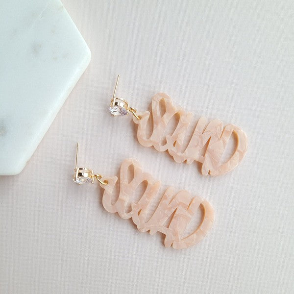 Two dangling earrings featuring "chaos" in peach-colored material, with exquisite hand-lettered calligraphy, are attached to gold posts adorned with clear stone accents. Perfectly named the "Bachelorette Squad," these earrings make a stunning bridal party gift and rest elegantly on a white surface.