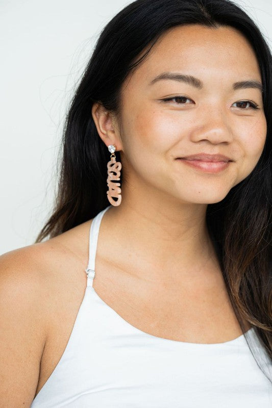 Two dangling earrings featuring "chaos" in peach-colored material, with exquisite hand-lettered calligraphy, are attached to gold posts adorned with clear stone accents. Perfectly named the "Bachelorette Squad," these earrings make a stunning bridal party gift and rest elegantly on a white surface.