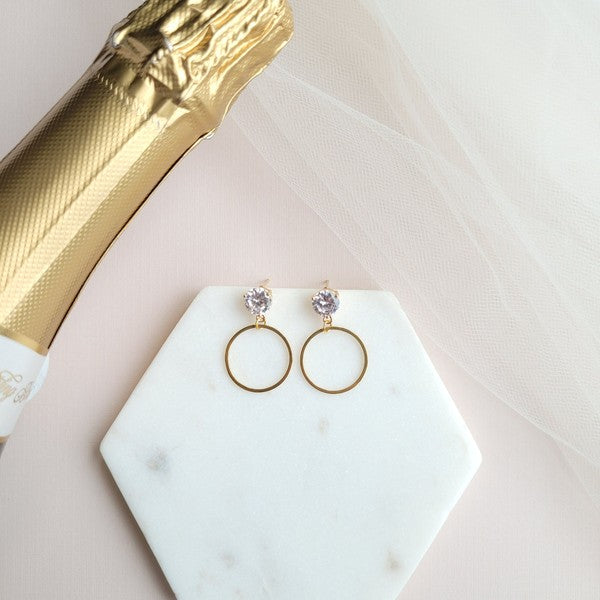 The I Do Earrings, adorned with gold and diamonds, rest elegantly on a hexagonal marble plate beside a champagne bottle, ready to catch the eye of wedding guests.