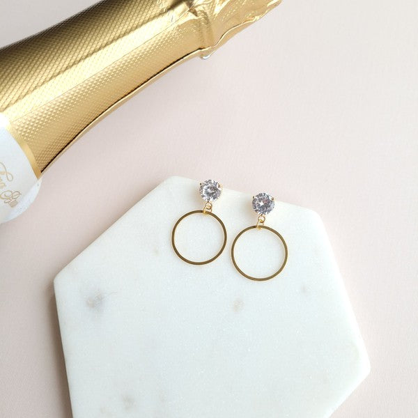 The I Do Earrings, adorned with gold and diamonds, rest elegantly on a hexagonal marble plate beside a champagne bottle, ready to catch the eye of wedding guests.