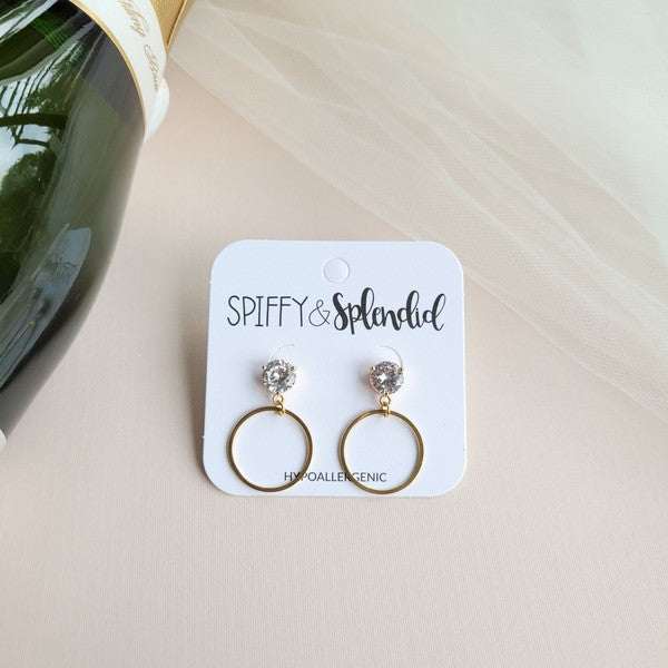 The I Do Earrings, adorned with gold and diamonds, rest elegantly on a hexagonal marble plate beside a champagne bottle, ready to catch the eye of wedding guests.