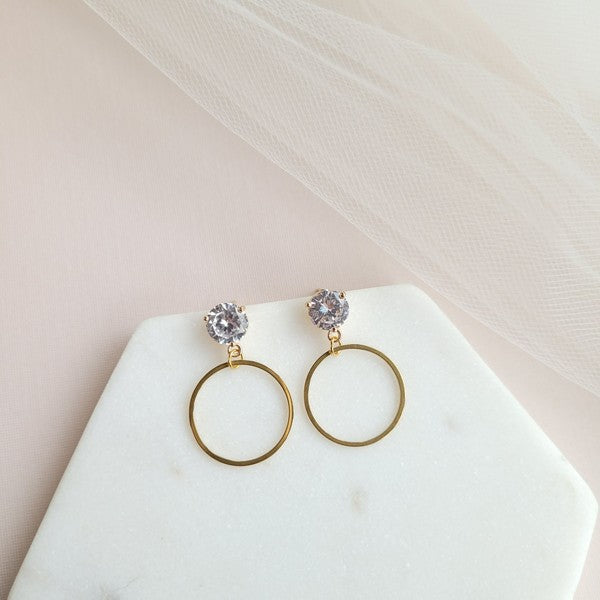 The I Do Earrings, adorned with gold and diamonds, rest elegantly on a hexagonal marble plate beside a champagne bottle, ready to catch the eye of wedding guests.