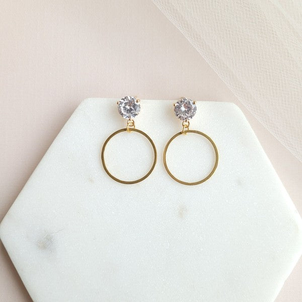 The I Do Earrings, adorned with gold and diamonds, rest elegantly on a hexagonal marble plate beside a champagne bottle, ready to catch the eye of wedding guests.