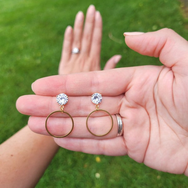 The I Do Earrings, adorned with gold and diamonds, rest elegantly on a hexagonal marble plate beside a champagne bottle, ready to catch the eye of wedding guests.