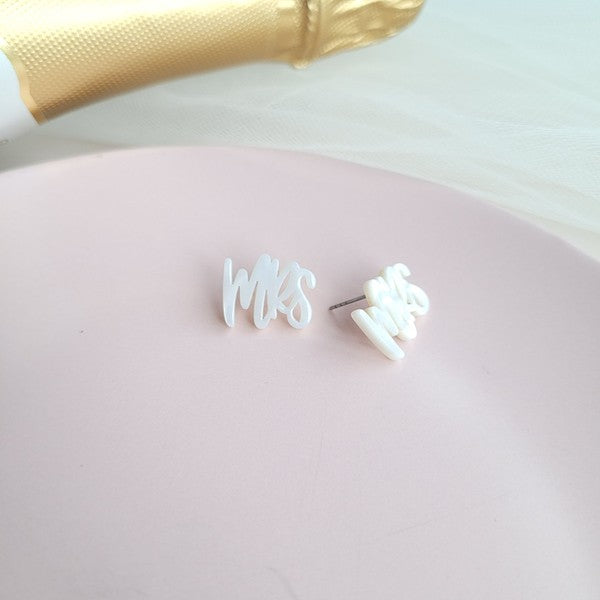 White earrings with custom hand-lettered calligraphy that spells "Mrs" are placed on a pink surface, with a hint of gold subtly in the background. An ideal choice for newlyweds, these are the elegant MRS Studs.