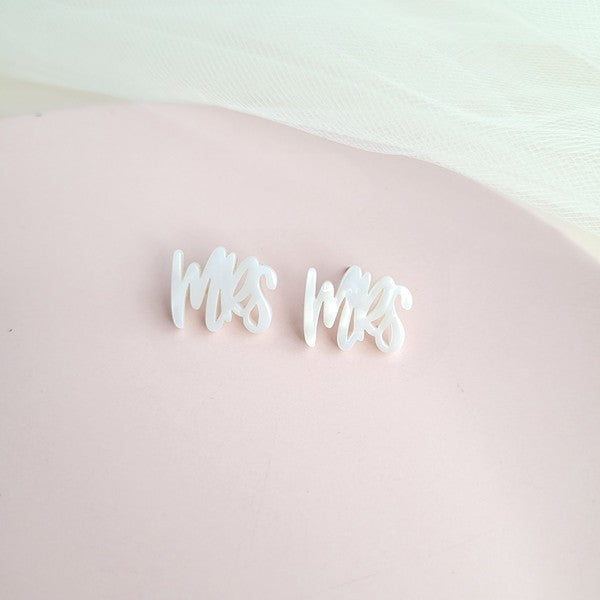 White earrings with custom hand-lettered calligraphy that spells "Mrs" are placed on a pink surface, with a hint of gold subtly in the background. An ideal choice for newlyweds, these are the elegant MRS Studs.