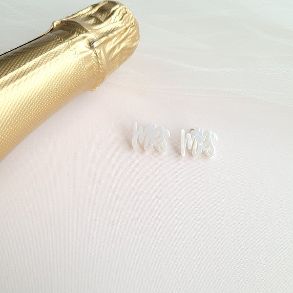 White earrings with custom hand-lettered calligraphy that spells "Mrs" are placed on a pink surface, with a hint of gold subtly in the background. An ideal choice for newlyweds, these are the elegant MRS Studs.