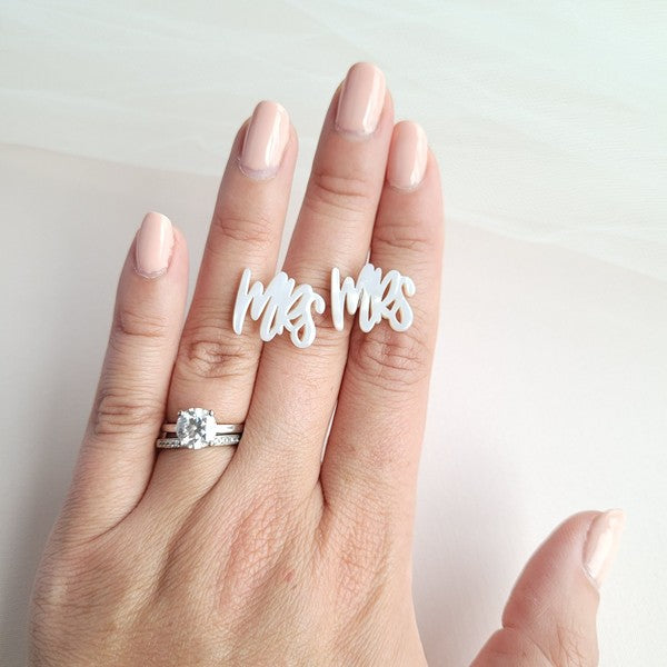 White earrings with custom hand-lettered calligraphy that spells "Mrs" are placed on a pink surface, with a hint of gold subtly in the background. An ideal choice for newlyweds, these are the elegant MRS Studs.