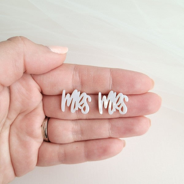 White earrings with custom hand-lettered calligraphy that spells "Mrs" are placed on a pink surface, with a hint of gold subtly in the background. An ideal choice for newlyweds, these are the elegant MRS Studs.