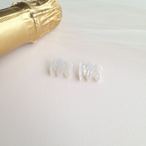 White earrings with custom hand-lettered calligraphy that spells "Mrs" are placed on a pink surface, with a hint of gold subtly in the background. An ideal choice for newlyweds, these are the elegant MRS Studs.