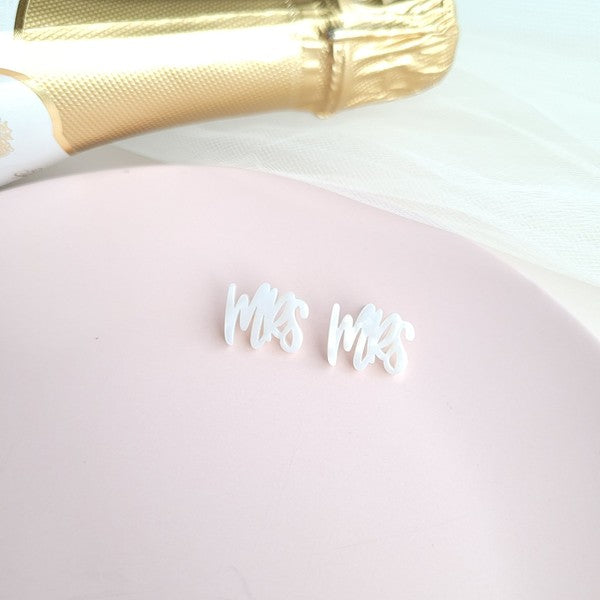 White earrings with custom hand-lettered calligraphy that spells "Mrs" are placed on a pink surface, with a hint of gold subtly in the background. An ideal choice for newlyweds, these are the elegant MRS Studs.