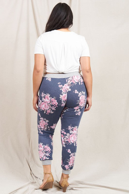 The individual, dressed in Floral Drawstring Joggers that are crafted in the United States and accented with a white drawstring, is paired with white sneakers and positioned against a white wall.