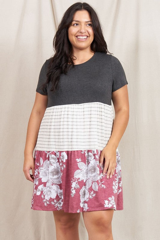 A woman dons the Short Sleeve Floral Tiered Midi Dress, showcasing a dark top, striped middle section, and a floral-patterned skirt. Made in the USA, this elegant dress creates a striking silhouette against its plain backdrop.