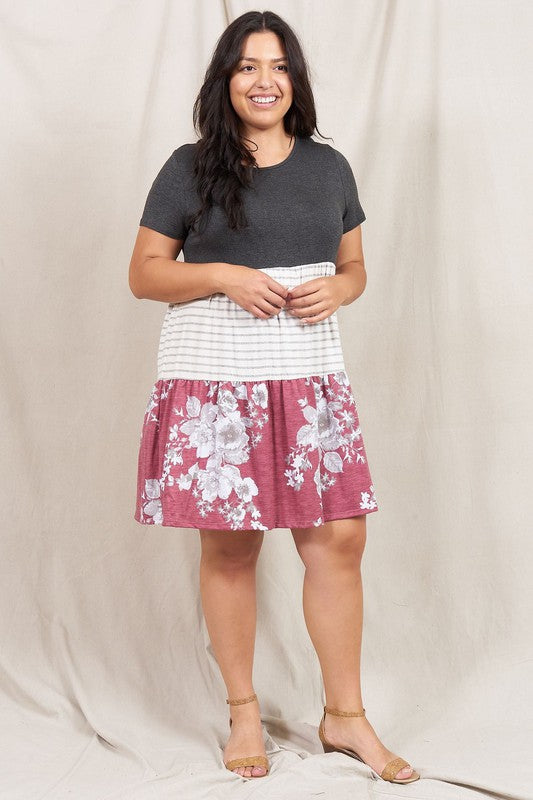 A woman dons the Short Sleeve Floral Tiered Midi Dress, showcasing a dark top, striped middle section, and a floral-patterned skirt. Made in the USA, this elegant dress creates a striking silhouette against its plain backdrop.