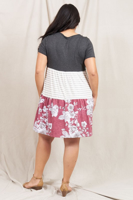 A woman dons the Short Sleeve Floral Tiered Midi Dress, showcasing a dark top, striped middle section, and a floral-patterned skirt. Made in the USA, this elegant dress creates a striking silhouette against its plain backdrop.