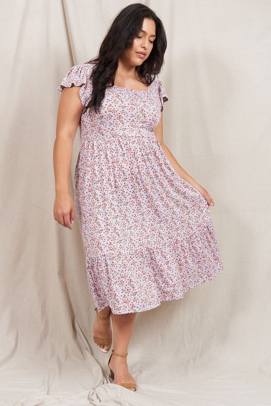 A woman wearing the Plus Flutter Sleeve Ditsy Floral Sundress paired with sandals stands in front of a plain fabric background.