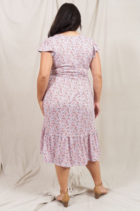 A woman wearing the Plus Flutter Sleeve Ditsy Floral Sundress paired with sandals stands in front of a plain fabric background.