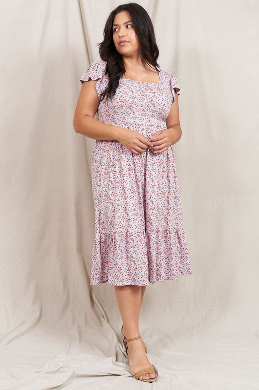 A woman wearing the Plus Flutter Sleeve Ditsy Floral Sundress paired with sandals stands in front of a plain fabric background.