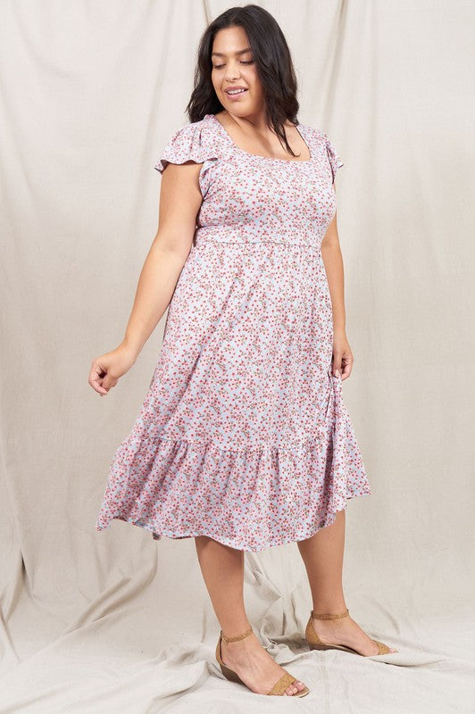 A woman wearing the Plus Flutter Sleeve Ditsy Floral Sundress paired with sandals stands in front of a plain fabric background.