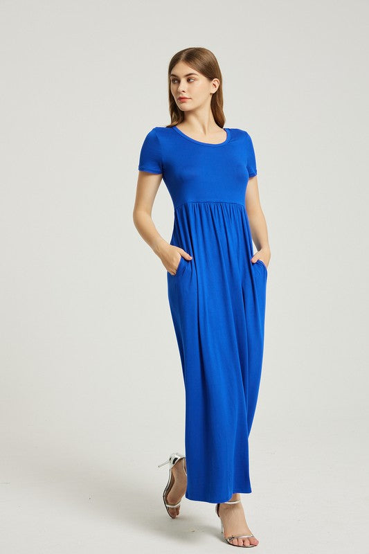 A woman in a blue Women's Summer Casual Maxi Dress With Pocket sits on a white chair. The short sleeve design complements her look as she gazes at the camera with a neutral expression.