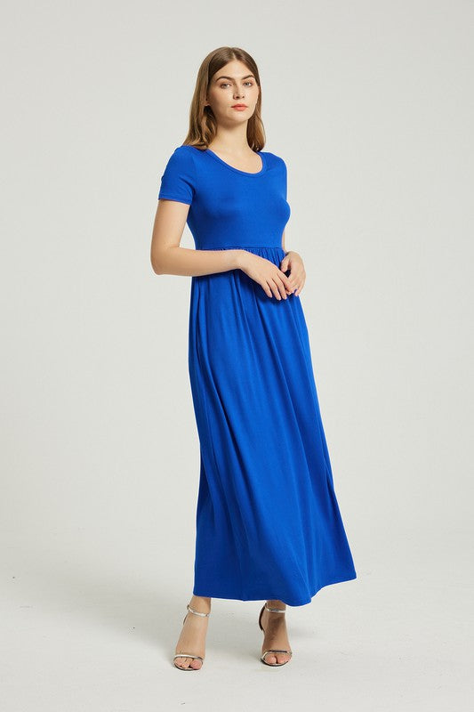 A woman in a blue Women's Summer Casual Maxi Dress With Pocket sits on a white chair. The short sleeve design complements her look as she gazes at the camera with a neutral expression.