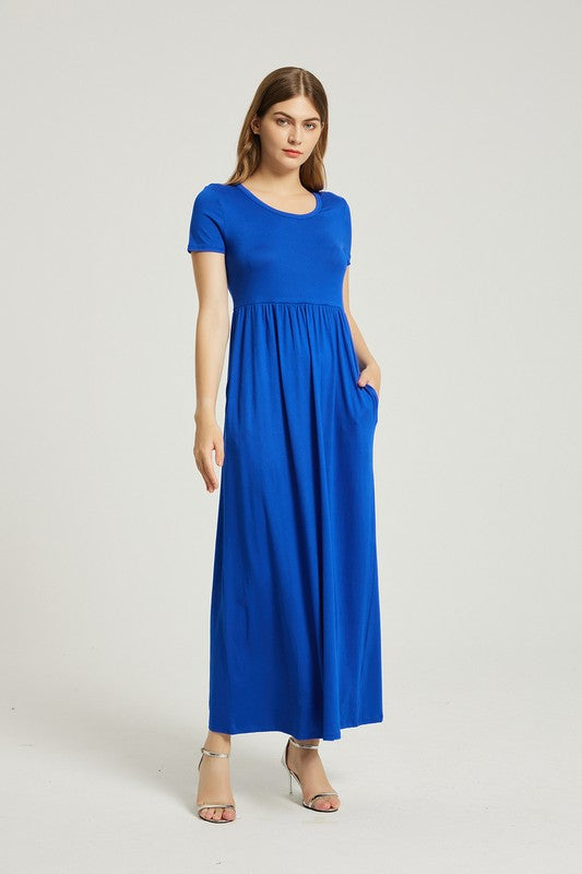 A woman in a blue Women's Summer Casual Maxi Dress With Pocket sits on a white chair. The short sleeve design complements her look as she gazes at the camera with a neutral expression.