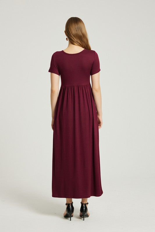 A woman in a Women's Summer Casual Maxi Dress With Pocket in burgundy sits on a wooden chair with one arm resting on the backrest. She wears black open-toe heels, and the background is a plain light gray. The stretch fabric of her dress enhances its comfort and style.