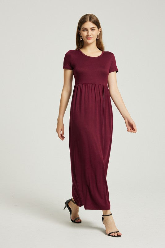 A woman in a Women's Summer Casual Maxi Dress With Pocket in burgundy sits on a wooden chair with one arm resting on the backrest. She wears black open-toe heels, and the background is a plain light gray. The stretch fabric of her dress enhances its comfort and style.