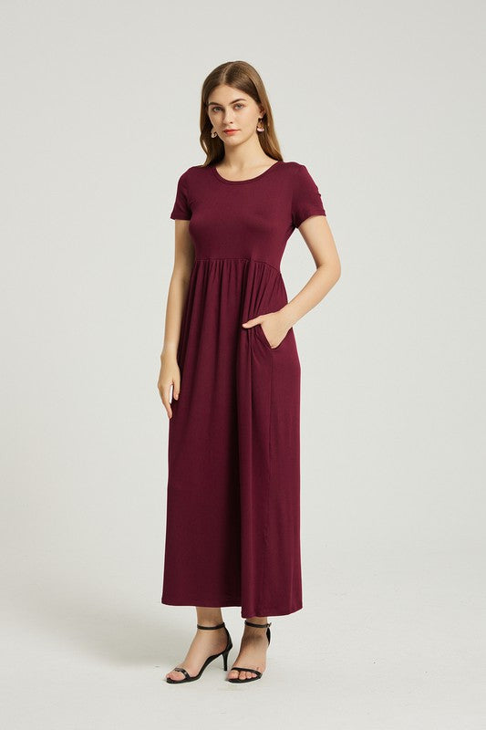 A woman in a Women's Summer Casual Maxi Dress With Pocket in burgundy sits on a wooden chair with one arm resting on the backrest. She wears black open-toe heels, and the background is a plain light gray. The stretch fabric of her dress enhances its comfort and style.