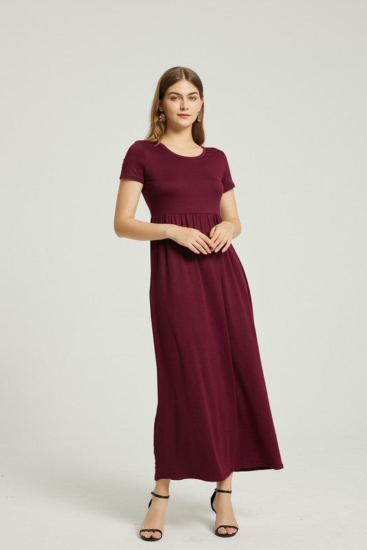 A woman in a Women's Summer Casual Maxi Dress With Pocket in burgundy sits on a wooden chair with one arm resting on the backrest. She wears black open-toe heels, and the background is a plain light gray. The stretch fabric of her dress enhances its comfort and style.