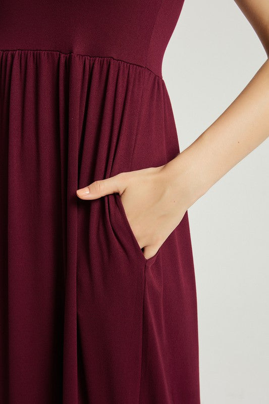 A woman in a Women's Summer Casual Maxi Dress With Pocket in burgundy sits on a wooden chair with one arm resting on the backrest. She wears black open-toe heels, and the background is a plain light gray. The stretch fabric of her dress enhances its comfort and style.