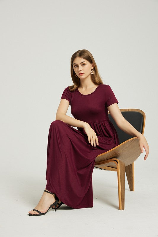 A woman in a burgundy Women's Summer Casual Maxi Dress With Pocket, featuring a Solid Pattern, sits on a wooden chair with one arm resting on the chair's backrest and her legs crossed. The background is plain and light-colored.