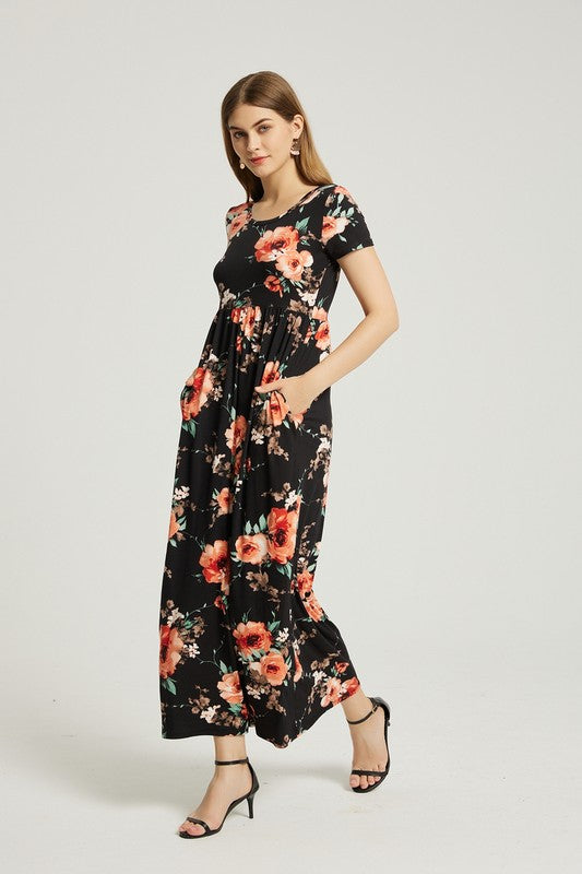 A woman wearing the Womens Summer Casual Floral Maxi Dress With Pocket in black floral is sitting on a modern wooden chair, resting her chin on her left hand and looking at the camera.