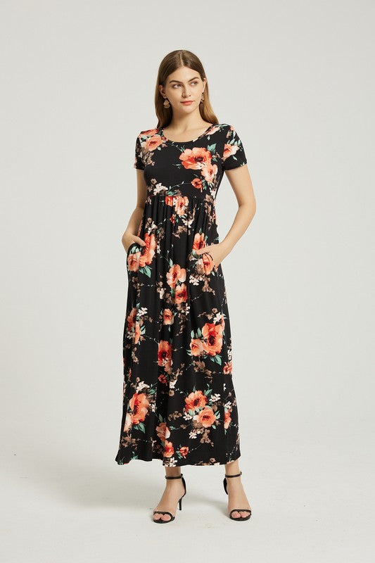 A woman wearing the Womens Summer Casual Floral Maxi Dress With Pocket in black floral is sitting on a modern wooden chair, resting her chin on her left hand and looking at the camera.