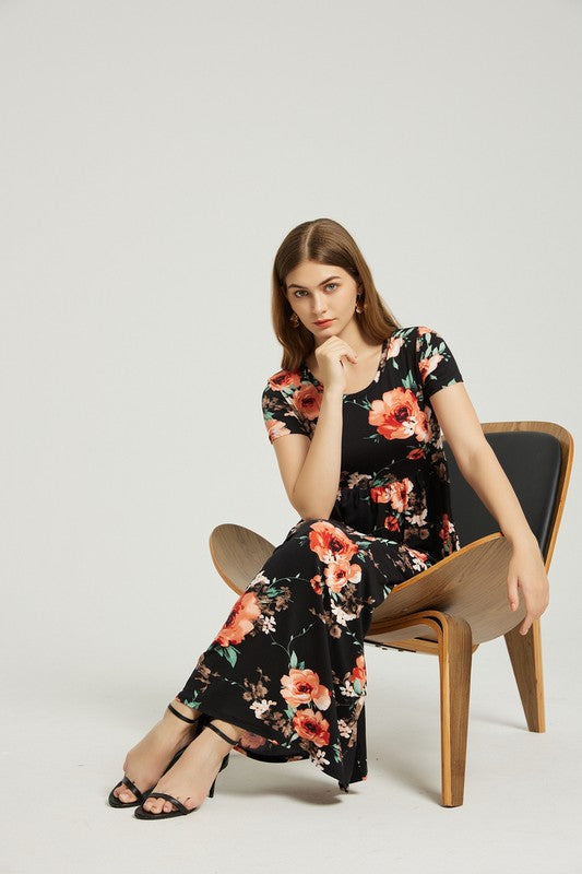 A woman sits on a wooden chair, clad in a black Women's Summer Casual Floral Maxi Dress With Pocket and matching shoes, with one hand resting on her chin and the other on her knee.