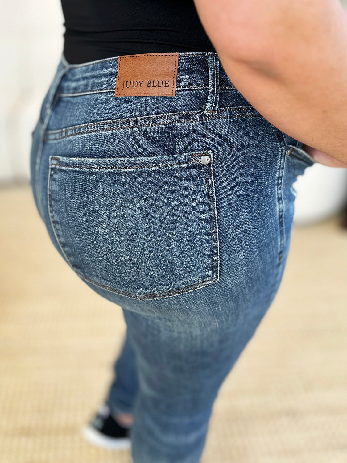 Person standing indoors wearing Judy Blue Full Size Mid Waist Distressed Slim Jeans, a black top, and black sneakers with white laces. There is a white sofa and a textured rug in the background.