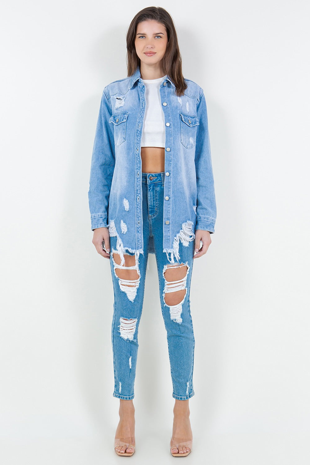 A woman stands against a plain background wearing the American Bazi Raw Hem Distressed Long Sleeve Denim Jacket over a white crop top and ripped jeans, adding an edgy flair to her wardrobe.